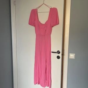 Rosa långklänning från Stradivarius - Säljer en rosa långklänning från Stradivarius med puffiga korta ärmar och snygg snörning i ryggen. Klänningen har en mjukt rynkad struktur och djup urringning både fram och bak. Perfekt för dig som vill sticka ut med färg och coola detaljer.