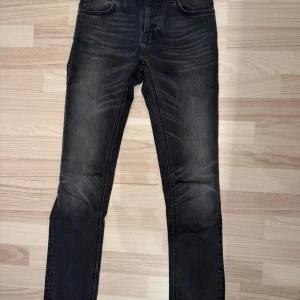 Svarta jeans från Nudie Jeans, W27 L30 - Snygga svarta jeans från Nudie Jeans med klassisk femficksdesign och coola slitningar. Jeansen har raka ben och subtila detaljer på bakfickorna. Passar dig som gillar en stilren look med lite edge. Materialet är slitstarkt denim och midjan är normal. Nyskick