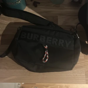 Svart axelväska från Burberry - Snygg svart axelväska från Burberry med bred axelrem och tydlig logga framtill. Väskan har en dragkedja med färgglad snodd och är tillverkad i slitstark syntet. Perfekt för dig som vill ha en stilren och praktisk väska med streetkänsla.
