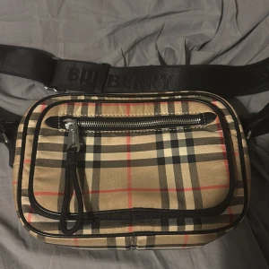 Burberry rutig axelväska med dragkedja - Snygg axelväska från Burberry med klassiskt beige, svart och rött rutmönster. Väskan har svarta detaljer i skinn, bred axelrem med Burberry-logga och flera fack med dragkedja. Perfekt storlek för det viktigaste och riktigt stilren design. FINNS EJ KVITTO!!!