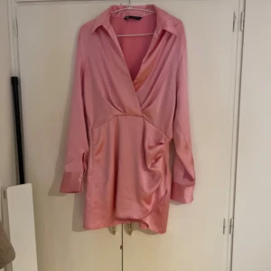Rosa omlottklänning från Zara - Snygg rosa kortklänning från Zara i satinliknande polyester. Klänningen har omlott-design med djup v-ringning, långa ärmar med breda manschetter och krage. Snygg drapering på sidan ger en cool och trendig look.