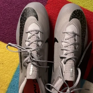 Nike Mercurial Air Zoom fotbollsskor elite Nike  - Säljer ett par grå Nike Mercurial Air Zoom fotbollsskor med svart swoosh och coola grafiska detaljer. Skorna har snörning, platt sula med dobbar och är tillverkade i syntetmaterial för bästa grepp och känsla på planen. Perfekta för dig som vill sticka ut på matchen!