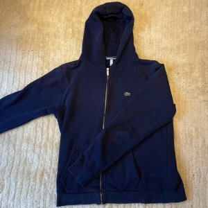 Marinblå hoodie från Lacoste - Snygg marinblå hoodie från Lacoste. Inte så använt och inget fel med den. Dragkedjan funkar helt, den har två fickor och en hood. Passade inte riktigt mig första gången ja köpte den några månader sen. Och tänkte sälja den eftersom de är i väldigt bra skick med.