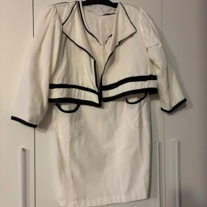 Vit kortklänning med svarta detaljer - Snygg vit kortklänning med svarta kantdetaljer och markerad krage. Klänningen har trekvartsärmar, två fickor framtill och en loose passform. Perfekt för dig som gillar kontraster och stilrena linjer.