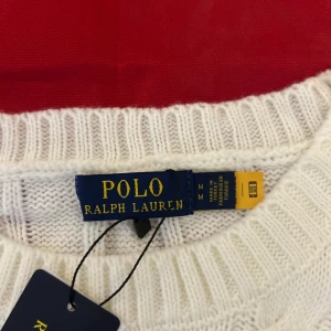 Vit kabelstickad tröja Polo Ralph Lauren - Vit kabelstickad tröja från Polo Ralph Lauren med klassisk rund halsringning och broderad logga på bröstet. Tröjan har långa ärmar och ribbade muddar för en snygg passform. Perfekt för dig som gillar stilrena och tidlösa plagg.