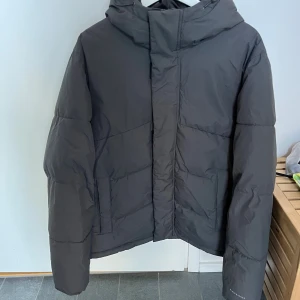 Svart pufferjacka från Jack & Jones - Säljer en svart pufferjacka från Jack & Jones i storlek M. Jackan har huva, raka linjer och diskret logga på ärmen. Perfekt för kalla dagar med sin vadderade design och klassiska look. Stängs med dragkedja och har två sidofickor.