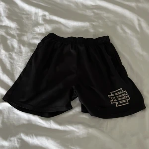 Svarta shorts från Eric Emanuel - Säljer ett par svarta shorts från Eric Emanuel med elastisk midja och logga broderad i vitt på ena benet. De är tillverkade i ett glansigt syntetmaterial och har en loose passform som är perfekt för träning eller chill dagar.