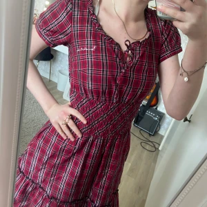 Rutig röd kortklänning från Bondelid - Supersöt röd och vit rutig kortklänning från Bondelid med smockad midja, volangkant nertill och knytning vid halsen. Puffiga korta ärmar och broderad text på bröstet. Osynliga fickor nertill.  Perfekt att ha på julen eller liknande 💯💋Använd 2 gånger.