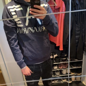 Marinblå hoodie från Armani - Snygg marinblå hoodie från Armani med stor vit logga och grafiska ränder på bröstet. Tröjan har huva med snörning och en praktisk magficka. Perfekt för dig som gillar streetwear och vill ha en cool look.