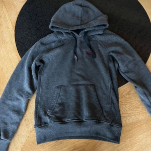Mörkgrå hoodie med röd logga - Mörkgrå hoodie från Swedemount med röd logga på bröstet. Tröjan har huva med snörning, stor magficka och ribbade muddar vid ärmslut och nederkant. Perfekt för chill eller träning, snygg och enkel stil.