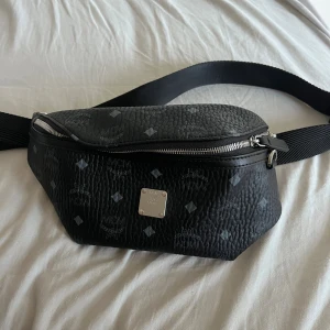 Svart magväska från MCM i skinn - Snygg svart magväska från MCM i präglat skinn med silverfärgade detaljer och dragkedjor. Väskan har MCM-logga och mönster över hela ytan samt justerbart band. Perfekt för att bära det viktigaste med stil.