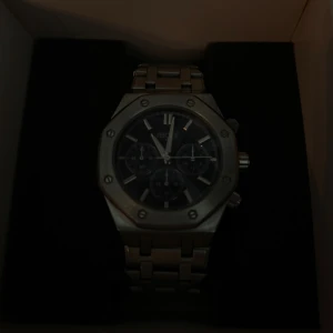 Regal silverfärgad kronografklocka - Stilren klocka från Regal med silverfärgad länk i metall och svart urtavla. Klockan har kronograf-funktion, markerade index och detaljerade visare. Boetten är åttakantig med synliga skruvar för en modern look. Perfekt accessoar för dig som gillar snygga detaljer.