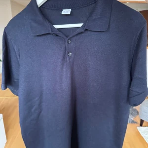 Mörkblå pikétröja från Lindex - Snygg mörkblå pikétröja från Lindex med klassisk krage och två knappar framtill. Tröjan har korta ärmar och är gjord i mjuk bomull som känns skön mot huden. Perfekt för dig som gillar enkel och stilren look. Passar storlek S!