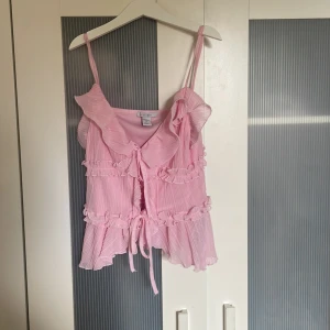 Rosa volangtop från Amisu - Säljer en ljusrosa topp från Amisu i storlek S. Toppen har tunna axelband, volangdetaljer och knyt framtill. Materialet är lätt och plisserat, vilket ger en flowy känsla. Perfekt för dig som gillar romantiska och söta vibes.