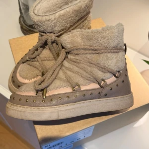 Beige boots med nitar och snörning - Säljer ett par beige boots från INUIKII med mjuk pälsimitation och grov snörning runt hela skon. Yttermaterialet är mocka och skinn, dekorerat med små runda nitar längs kanten. Perfekta för kalla dagar med sin fluffiga insida och platta sula. Modellen heter curly rock. Köpta förra året för 3999kr men sparsamt använda så fint skick.