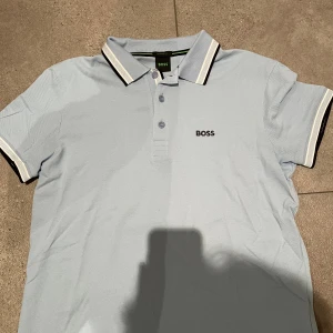 Ljusblå pikétröja från BOSS - Snygg ljusblå pikétröja från BOSS med klassisk krage och svarta/vita ränder på krage och ärmslut. Regular fit och tillverkad i mjuk bomull. BOSS-logga broderad på bröstet. Perfekt för dig som gillar stilrena och sportiga vibes.