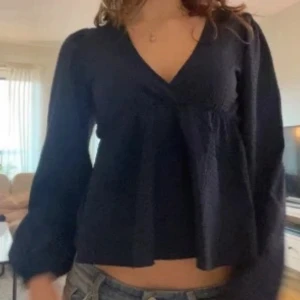 Mörkblå omlottblus med puffärm - Säljer en mörkblå blus från Nelly med omlott-design och v-ringning. Blusen har långa puffärmar och är tillverkad i ett lätt, strukturerat tyg som ger en cool känsla. Perfekt att styla med jeans eller kjol för en trendig look.