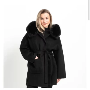 Svart kappa med fuskpäls och bälte - Stilren svart kappa med bred krage i fuskpäls och knytskärp i midjan. Jackan har stora fickor framtill och en rak, loose passform som ger en cool och modern vibe. Perfekt för kalla dagar när du vill vara både varm och snygg.