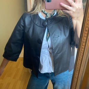 Svart skinnjacka med stora knappar - Snygg svart skinnjacka från InWear med stora klädda knappar och trekvartsärm. Jackan har en rundad krage och en loose passform som ger en cool och retro vibe. Perfekt att slänga över en skjorta eller t-shirt för en stilren look.