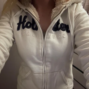 Vit hoodie från Hollister med foder - Mysig vit hoodie från Hollister med fluffigt foder i huvan och insidan. Tröjan har dragkedja, broderad logga i mörkblått och snörning i huvan. Perfekt för kyliga dagar och har två fickor framtill.