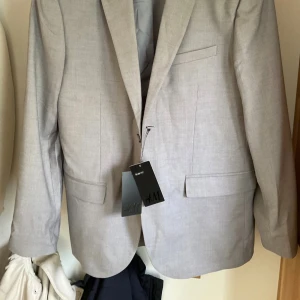 Grå kavaj stl 52 ullmix Nyskick - Gråmelange H&M kavaj i slim fit modell. Kavajen har en klassisk enkelknäppt. Den har även fickor med lock och en bröstficka. Stl 52, har lagt ner ärmarna ca 2 cm. Polyester viscose och 7 % ull cool max kvalitet. Nyskick. Lapparna ej kvar som på bild.  Nypris: 799kr