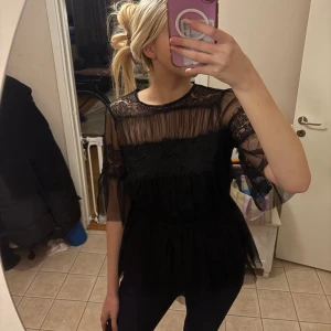 Svart blus i mesh och spets Happy Holly - Supersnygg svart blus från Happy Holly med transparent mesh och spetsdetaljer. Blusen har trekvartsärmar, volang och en lätt utsvängd passform. Perfekt för dig som gillar en trendig och feminin stil.