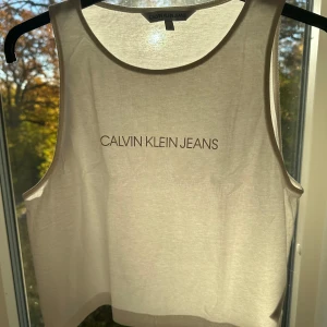 Vit linnetopp från Calvin Klein Jeans - Säljer en fräsch vit linnetopp från Calvin Klein Jeans med tryckt logga framtill. Toppen har rund halsringning och är ärmlös, perfekt för varma dagar. Materialet är mjuk bomull som känns skönt mot huden. Enkel och clean design som funkar till jeans eller shorts.