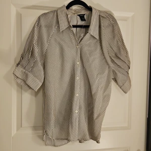 Randig skjorta från Lindex, M - Snygg skjorta från Lindex i storlek M med svartvita ränder. Skjortan har klassisk krage, knappar framtill och korta ärmar med uppvik. Materialet är mjukt och skönt, tillverkat av 85% lyocell och 15% polyester. Perfekt för dig som gillar stilrena och moderna plagg.