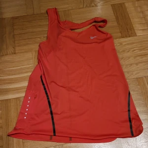 Nike Dri-Fit röd linnetopp S - Sportig röd linnetopp från Nike med Dri-Fit-teknologi. Toppen har svarta ränder, reflekterande prickar på sidan och brottarrygg. Perfekt för träning och gjord i ett lätt syntetmaterial som andas.