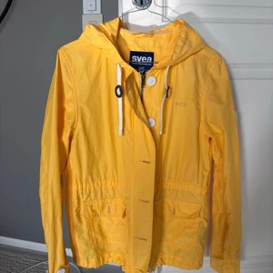 vindjacka med huva Svea XS/S - Gul vindjacka från Svea med huva, vita snören och dragkedja framtill. Jackan har två fickor med lock, resår i midjan och en patch med amerikansk flagga på ena ärmen. Perfekt för blåsiga dagar och snygg till jeans.