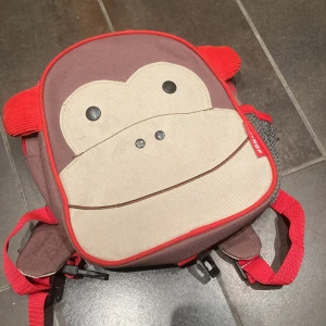 Ryggsäck med apa från Skip Hop - Söt ryggsäck från Skip Hop med apansikte framtill. Väskan är brun, beige och röd med röda detaljer och dragkedja. Tillverkad i slitstark polyester med meshficka på sidan och justerbara axelband. Perfekt för skola eller utflykt.