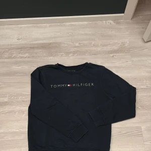 Mörkblå Tommy Hilfiger sweatshirt - Snygg mörkblå sweatshirt från Tommy Hilfiger med klassisk logga tryckt på bröstet. Tröjan har rund halsringning, långa ärmar och ribbade muddar. Perfekt för dig som gillar stilrena och bekväma plagg med ikonisk branding.