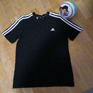 Svart Adidas t-shirt med vita ränder - Svart t-shirt från Adidas med klassiska vita ränder längs axlarna och ärmarna. Adidas-logga på bröstet. Skön passform och perfekt för fotboll eller träning. Tillverkad i mjukt material som andas.