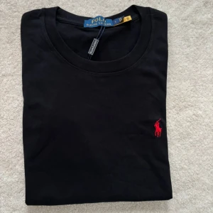 Svart t-shirt från Polo Ralph Lauren - Svart t-shirt från Polo Ralph Lauren i mjuk bomull. Klassisk rund hals och korta ärmar. På bröstet finns den ikoniska röda broderade Polo-loggan. Enkel och stilren design som funkar till allt. Snygg passform och skön kvalitet.
