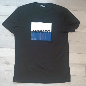 Svart t-shirt Antony Morato S - Svart t-shirt från Antony Morato i storlek S. Framsidan har en stor vit och blå fyrkantig print med texten 'MORATO' i svart. Klassisk passform med rund hals och korta ärmar. Tillverkad i mjuk bomull för en skön känsla. ALDRIG ANVÄND! 