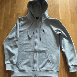 Grå hoodie från Polo Ralph Lauren - Snygg grå hoodie från Polo Ralph Lauren med dragkedja och huva. Klassisk logga broderad på bröstet, två fickor fram och ribbade muddar. Perfekt för chill dagar och enkel att matcha med jeans eller joggers.