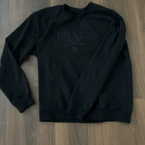Svart sweatshirt från Prada Milano - Säljer en svart sweatshirt från Prada Milano med broderad logga framtill. Tröjan har rund halsringning och långa ärmar. Tillverkad i mjukt bomullsmaterial som känns skönt mot huden. Perfekt för dig som gillar stilrena och exklusiva plagg.