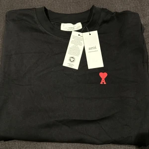 Svart t-shirt från Ami Paris M - Svart t-shirt från Ami Paris i mjuk bomull. Klassisk passform med korta ärmar och rund hals. På bröstet finns Ami:s ikoniska röda hjärta med A-broderi. Diskret AMI-broderi i nacken. Perfekt basic med snygg detalj.