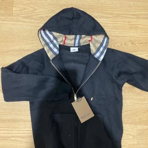Burberry hoodie (helt ny+gratis frakt) - Svart hoodie från Burberry med klassiskt rutigt foder i huvan, guldfärgad dragkedja och broderad logga på bröstet. Tröjan har långa ärmar, fickor framtill och är tillverkad i mjuk bomull. Perfekt för dig som vill ha en stilren och lyxig look.