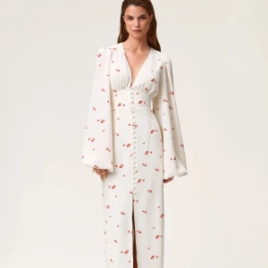 Vit långklänning med blommigt mönster - Superfin vit långklänning från Adoore med små röda blommor, djup v-ringning och puffiga långärmar. Klänningen har knappar framtill och markerad midja som ger en snygg siluett. Perfekt för sommarens alla tillfällen!