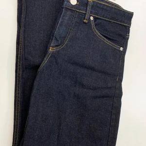 Mörkblå jeansbyxor från Design by Si XS  - Snygga mörkblå jeansbyxor från Design by Si i storlek XS med klassisk femficksdesign och kontrastsömmar i gult. Jeansen har en rak passform och är tillverkade i slitstarkt denimmaterial. Perfekt för dig som gillar en enkel och stilren look. Midja: 35,5 cm, Innerbenslängd: 76 cm, Längd från midja: 102 cm. 