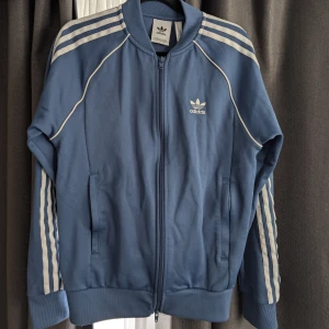 Blå Adidas track jacket med vita ränder - Säljer en blå Adidas trackjacket med klassiska vita ränder längs ärmarna och broderad logga på bröstet. Jackan har dragkedja framtill, ribbade muddar och två fickor. Perfekt för en sportig och avslappnad stil.