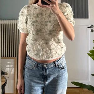 Blommig blus från Zara i ljusbeige - Superfin blus från Zara i ljusbeige med stora 3D-blommor och broderade detaljer. Puffiga korta ärmar och croppad passform ger en trendig vibe. Materialet är stickad med inslag av spets och broderi, perfekt för dig som gillar unika plagg.