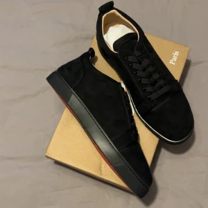 Christian Louboutin svarta sneakers - Snygga svarta sneakers från Christian Louboutin i mocka med klassisk röd sula och diskreta metalldetaljer på sidan. Skorna har rund tå, svarta skosnören och en stilren design som passar till allt. Kommer med originalkartong och tillbehör.