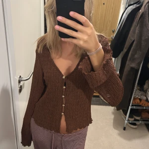 Brun blus med v-ringning och knappar - Säljer en brun blus med v-ringning och dekorativa pärlknappar framtill. Toppen har långärmade, lätt vågiga ärmar och ett ribbat, krusigt tyg som ger en cool struktur. Perfekt för dig som gillar unika detaljer och jordnära färger.