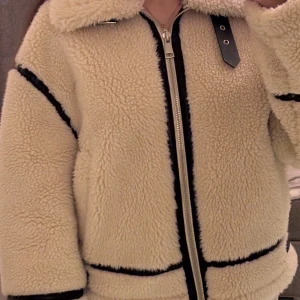 Beige teddyjacka från Zara  med svarta detaljer - Säljer en beige teddyjacka med svarta kontrastsömmar och dragkedja framtill. Jackan har stor krage, bälte vid midjan och muddar samt coola detaljer i svart läderimitation. Perfekt för dig som vill ha en mysig och trendig look i höst och vinter. Har andvänt den cirka 4 gånger 