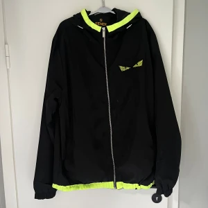 Svart vindjacka från Fendi med neon - Svart vindjacka från Fendi med huva och dragkedja framtill. Jackan har neonfärgade detaljer vid huva och nederkant samt ett coolt gult ögonmotiv på bröstet. Lätt och sportig känsla, perfekt för dig som gillar streetstyle.