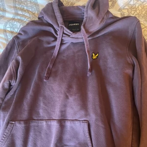 Lila hoodie från Lyle & Scott - Säljer en lila hoodie från Lyle & Scott med klassisk huva och dragsnören. Tröjan har en stor magficka och den ikoniska gula loggan på bröstet. Perfekt för dig som gillar en avslappnad och snygg stil.