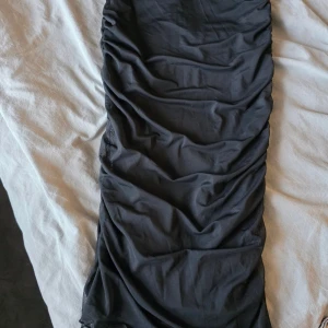 Svart långkjol från Gina Tricot - Svart långkjol från Gina Tricot med snygga rynkade detaljer längs sidorna. Kjolen har en smal passform och är tillverkad i ett mjukt, stretchigt polyestertyg som sitter bekvämt. Perfekt för dig som gillar en stilren och trendig look.