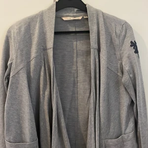 Grå kofta från House of Lola XS - Säljer en ljusgrå, mjuk och skön kofta i sweatshirt tyg från House of Lola i storlek XS. Koftan har öppen front, långa ärmar och två stora fickor framtill. Snygg broderad House of Lola älva på ena ärmen. Längre i fram, lite som en hängande sjal. House of Lola XS motsvarar normal S. 94% bomull 6% elastan 